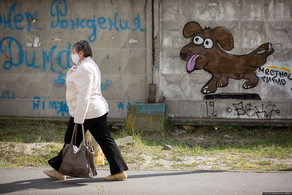 Жінка у медичній масці йде з торбами продуктів додому з магазину повз паркан зі смішним графіті. Фото - Андрій Дубчак