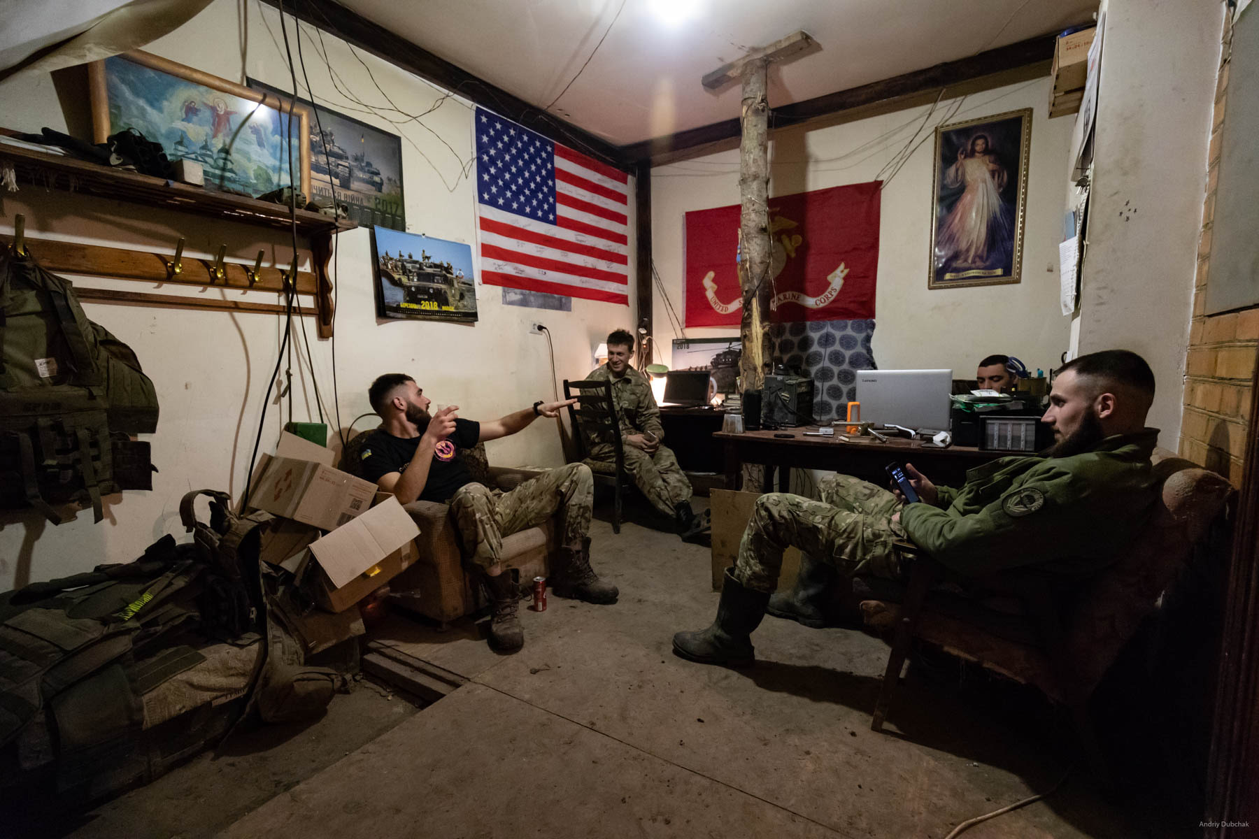 Marines converse in one of the basements in Vodyane. In the background, there is a portion of the US flag can be seen, donated by the American Marines to the Ukrainian ones, during joint exercises. The photo was taken in March, during our next visit in June, this house was gone. The enemy broke it by his mortars. Fortunately, the first mine that fell next to the house gave time for everyone to jump into the basement. Vodyane, March 2018.